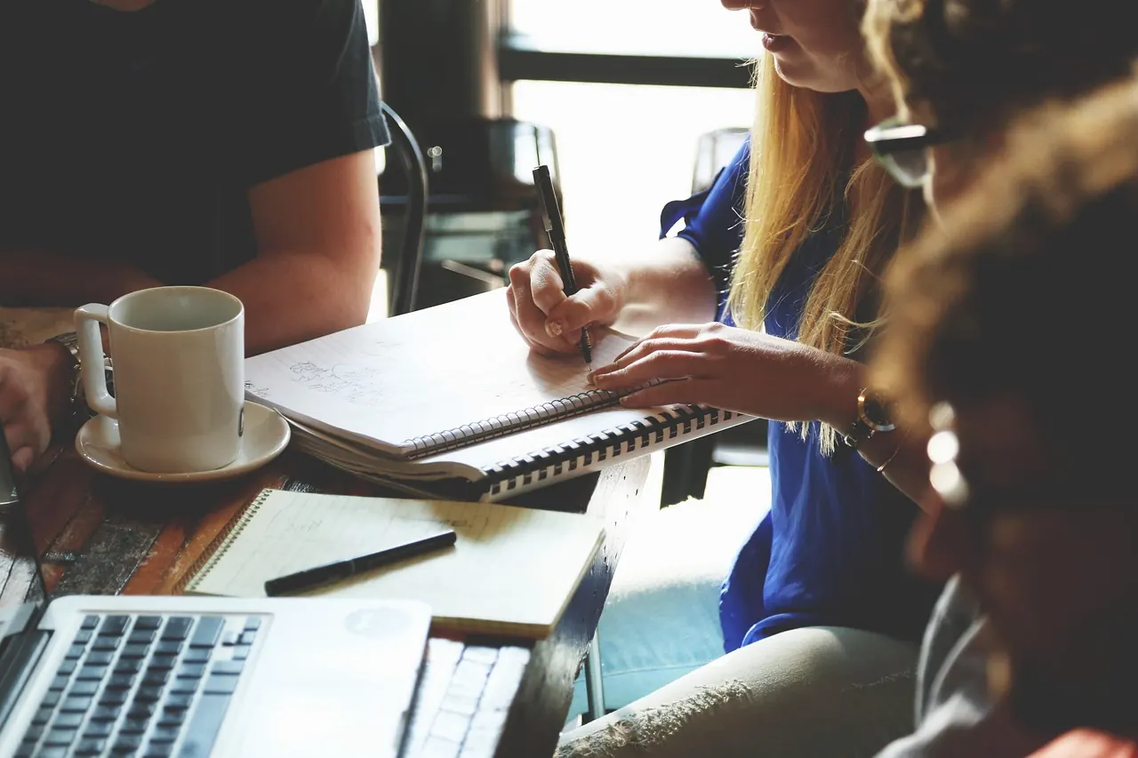 Três pessoas revisando anotações num caderno durante reunião, com café e notebook na mesa